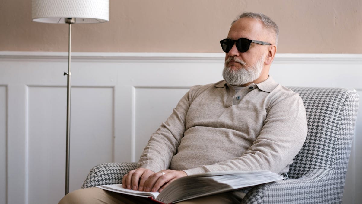 Senior man with sunglasses reading a braille book in a cozy modern interior.